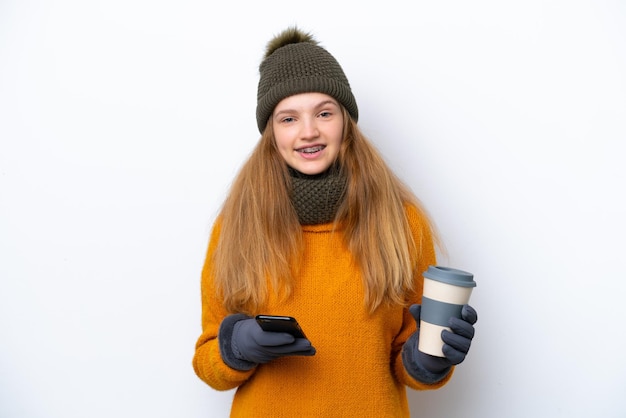 Teenager Russian girl wearing winter jacket isolated on white background holding coffee to take away and a mobile