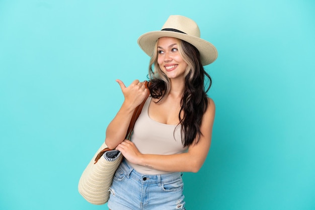 Teenager Russian girl with pamel and beach bag isolated on blue background pointing to the side to present a product