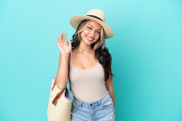 Teenager Russian girl with pamel and beach bag isolated on blue background saluting with hand with happy expression