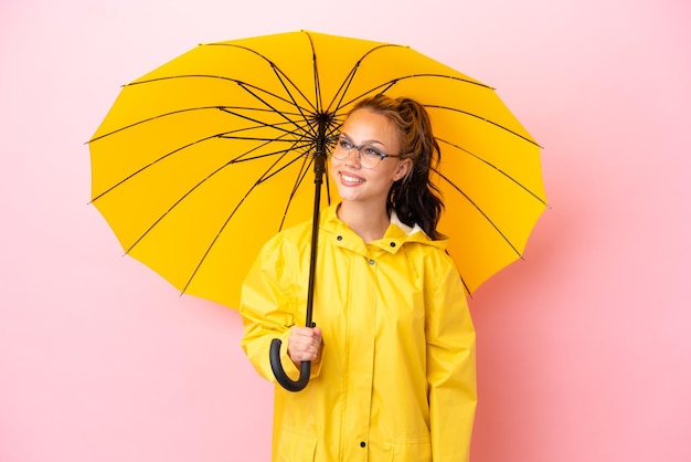 Teenager Russian girl with rainproof coat and umbrella isolated on pink background looking side