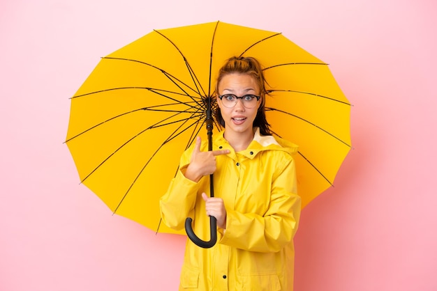 Teenager Russian girl with rainproof coat and umbrella isolated on pink background with surprise facial expression