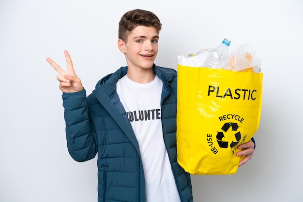 Teenager Russian holding a bag full of plastic bottles to recycle on white background smiling and showing victory sign