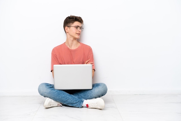 Teenager Russian man holding pc sitting on the floor isolated on white background looking side