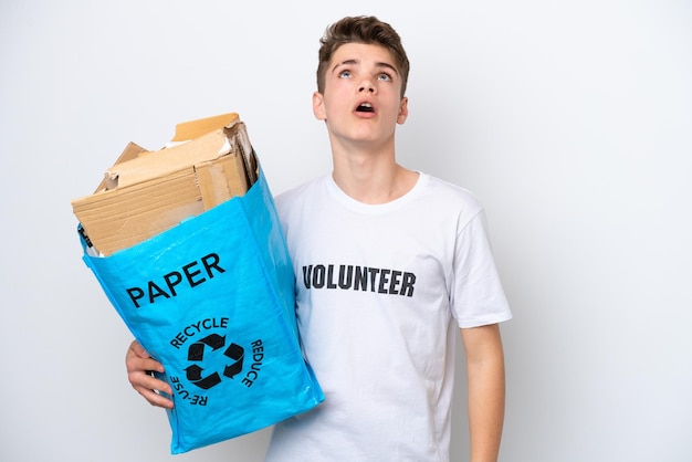 Teenager Russian man holding a recycling bag full of paper to recycle isolated on white background looking up and with surprised expression