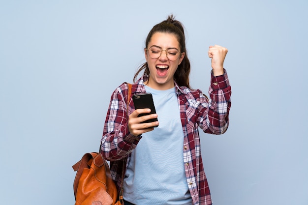 Teenager student girl over isolated blue wall with phone in victory position