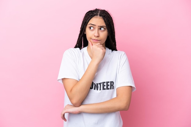 Teenager volunteer girl with braids isolated on pink background having doubts and thinking