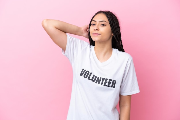 Teenager volunteer girl with braids isolated on pink background having doubts