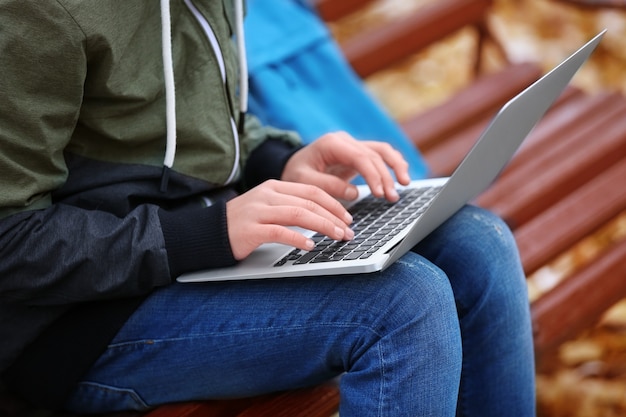 Teenager with laptop sitting on bench in autumn park, close up