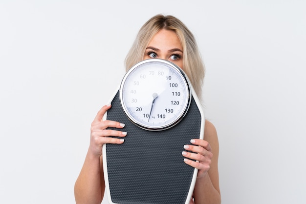 Teenager woman over isolated white wall with weighing machine and hiding behind it