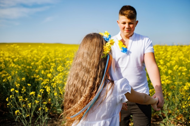 Teenagers brother and sister with Ukrainian wreath with on head in rapeseed field under blue sky