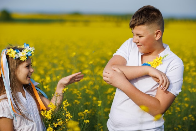 Teenagers brother and sister with Ukrainian wreath with on head in rapeseed field under blue sky