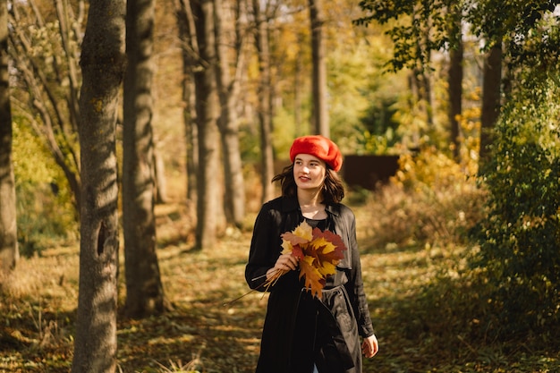 A teengirl in a red beret with a bouquet of autumn leaves in her hands walks through the forest