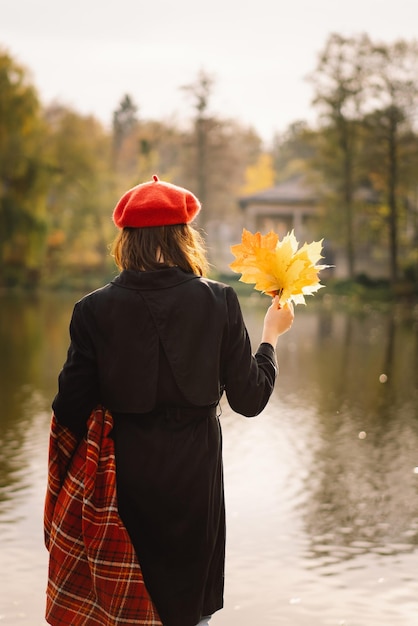 A Teengirl in a red beret with a bouquet of autumn leaves in her hands walks on wooden pontoon Autumn mood Autumn concept Autumn day Back view