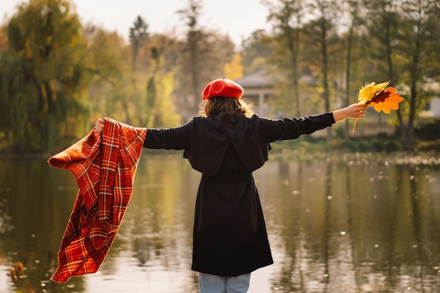 A Teengirl in a red beret with a bouquet of autumn leaves in her hands walks on wooden pontoon Autumn mood Autumn concept Autumn day Back view