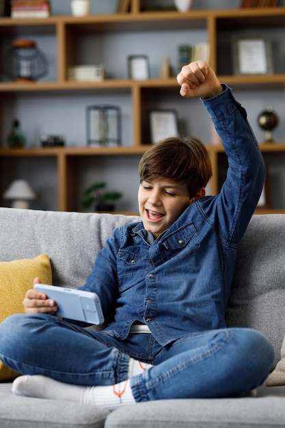 Teens and gaming addiction Young boy playing video game on smartphone sitting on sofa at home Cute teenager celebrating victory in online game on mobile phone Spending time at home leisure relax