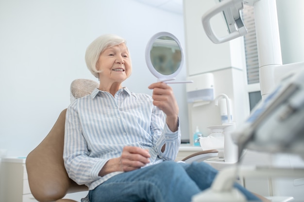 Teeth after restoration. Senior woman sitting at the dentists office and looking in the mirror after teeth restoration