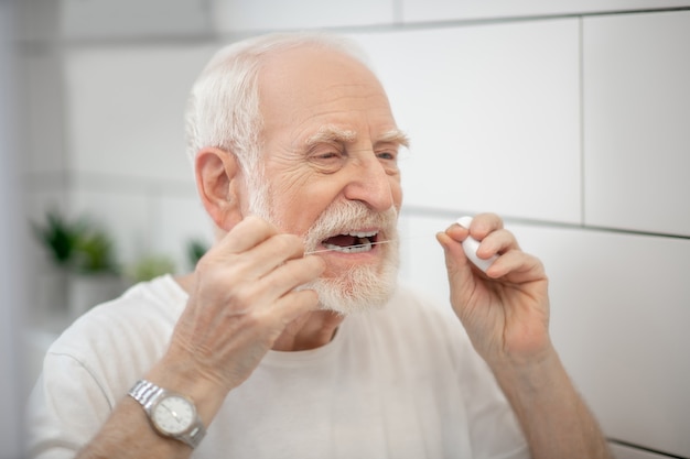 Teeth cleaning. Gray-haired man in white tshirt cleaning teeth with a floss