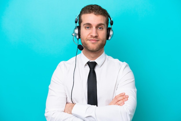 Telemarketer caucasian man working with a headset isolated on blue background keeping the arms crossed in frontal position