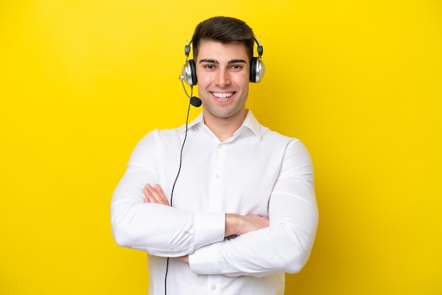 Telemarketer caucasian man working with a headset isolated on yellow background keeping the arms crossed in frontal position