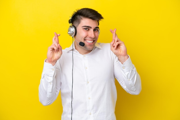 Telemarketer caucasian man working with a headset isolated on yellow background with fingers crossing