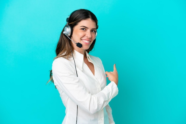 Telemarketer Italian woman working with a headset isolated on blue background pointing back