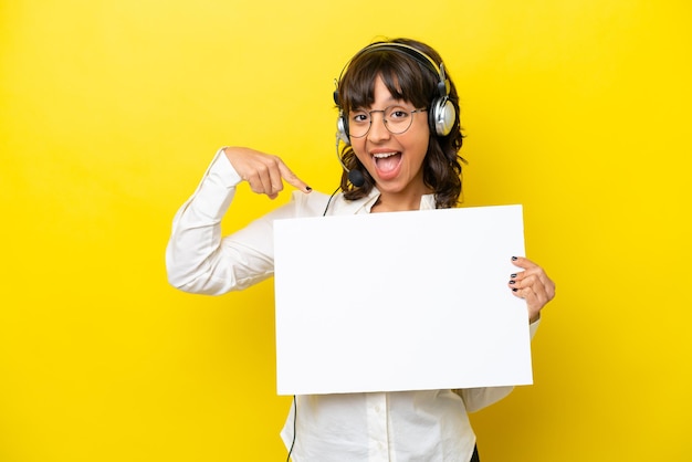 Telemarketer latin woman working with a headset isolated on yellow background holding an empty placard with happy expression and pointing it