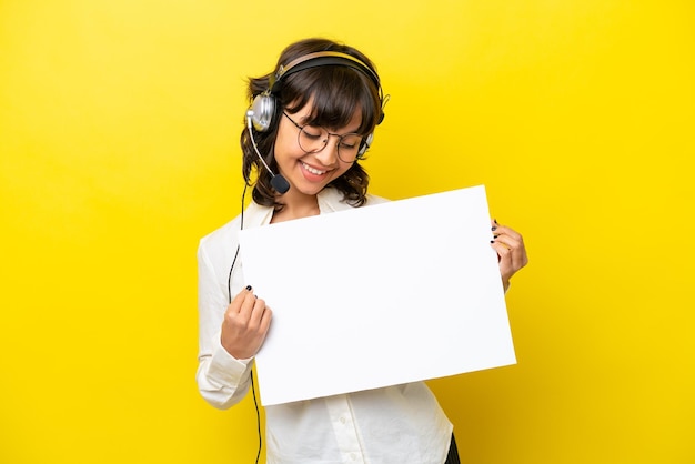 Telemarketer latin woman working with a headset isolated on yellow background holding an empty placard with happy expression