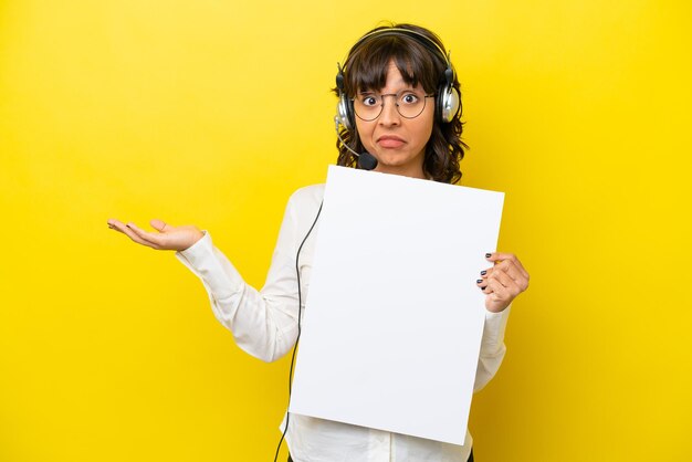 Telemarketer latin woman working with a headset isolated on yellow background holding an empty placard with surprised expression