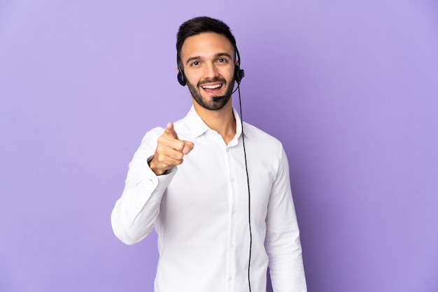 Telemarketer man working with a headset isolated on purple background surprised and pointing front