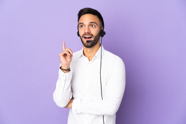 Telemarketer man working with a headset isolated on purple background thinking an idea pointing the finger up