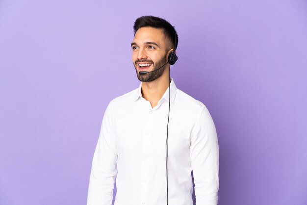 Telemarketer man working with a headset isolated on purple wall looking side