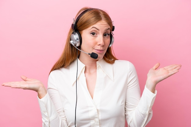 Telemarketer redhead woman working with a headset isolated on pink background having doubts while raising hands