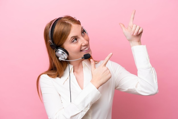 Telemarketer redhead woman working with a headset isolated on pink background pointing with the index finger a great idea