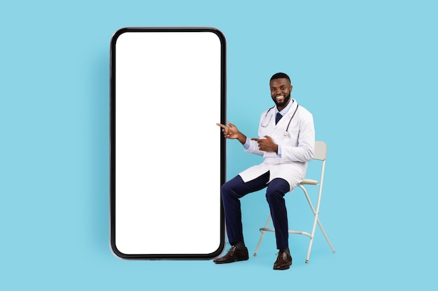 Telemedicine concept friendly black doctor sitting on chair near big blank smartphone