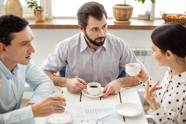 Telling news. Good-looking content bearded man drinking coffee and looking at a dark-haired woman holding a cup and talking