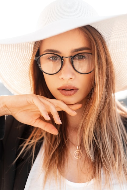 Tender beautiful young woman with pretty sexy lips with amazing eyes in youth trendy in fashionable hat in black blazer enjoys bright sunlight outdoors. Portrait charming girl on street in sunny day.