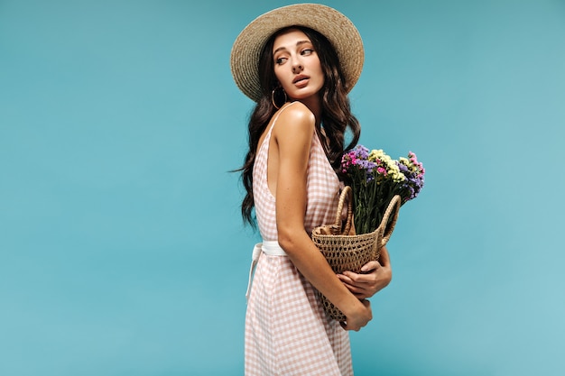 Tender charming woman with black wavy hair and earrings in stylish hat and summer dress posing with flowers on blue wall