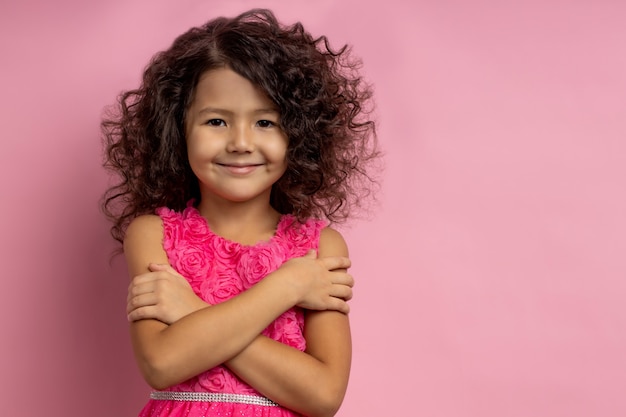 Tender cute smiling little girl with curly dark hair, wearing dress, hugging herself, looking with happy expression, posing with crossed hands, isolated with copy space.