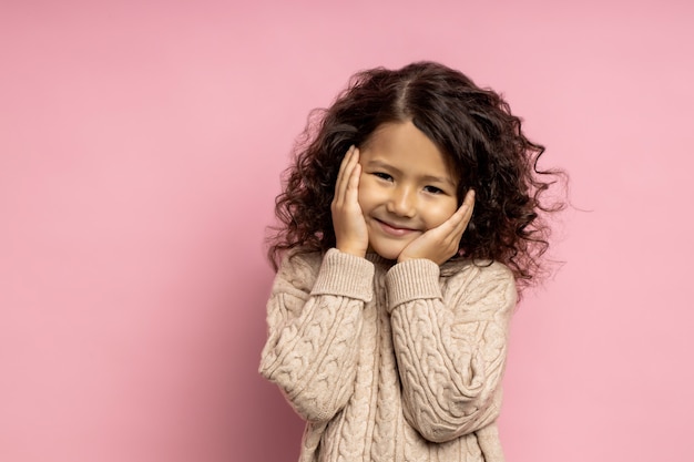 Tender dreamy shy small girl with kinky dark hair, touching cheeks with palms, having happy expression, wearing beige sweater, cute smiling, posing over pink wall.