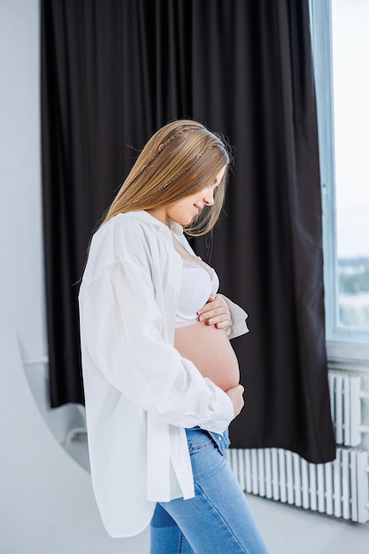 A tender young pregnant woman in a white shirt and jeans hugs her belly Pregnant happy woman on a white background