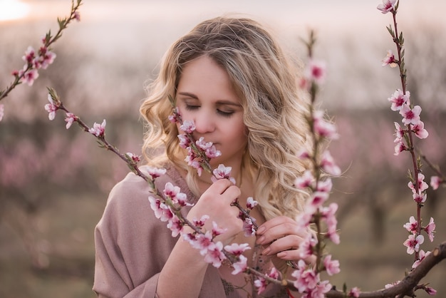 Tender young woman with beautiful curly blonde hair, with clean face, natural makeup, stands in blooming gardens, holds twig with pink inflorescences