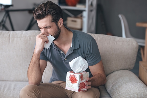 Terrible allergy. Frustrated handsome young man sneezing and using tissue while sitting on the couch at home