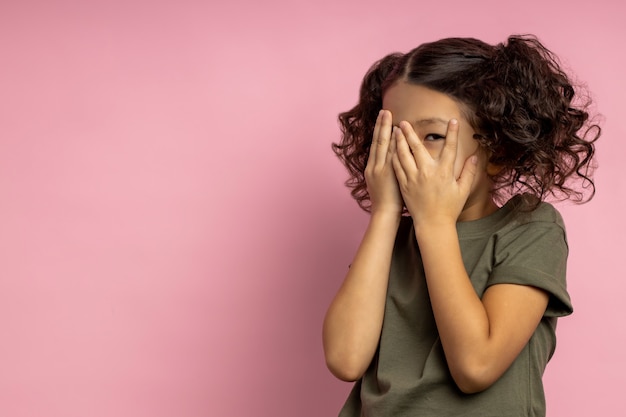 Terrified asian girl with curly hair, in khaki t shirt, covering face with both palms, hiding, peeking through fingers, watching scary cartoon. Human emotions. Copy space.