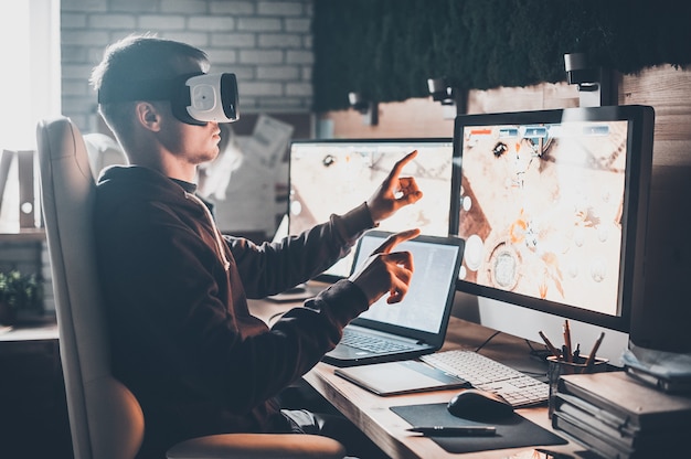 Testing games. Young man wearing virtual reality headset and gesturing while sitting at his desk in creative office