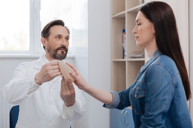 Testing the sample. Graceful thoughtful pretty woman paying a visit to plastic surgeon and learning about the procedure while he explaining her the specifics