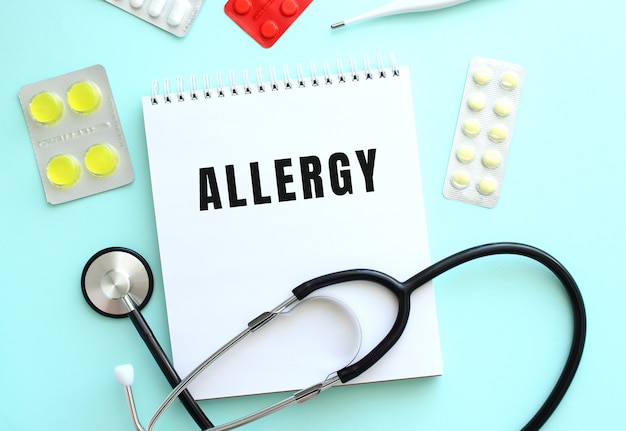 The text ALLERGY is written on a white notepad that lies next to the stethoscope and pills on a blue background.