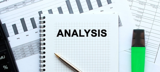 Text ANALYSIS on the page of a notepad lying on financial charts on the office desk. Near the calculator and marker.