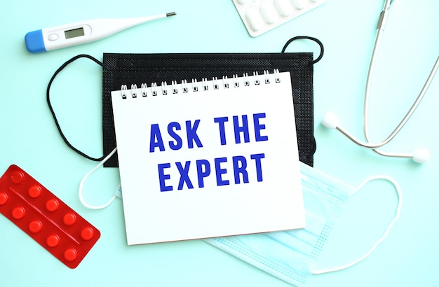 The text ASK THE EXPERT is written in a notebook that lies on a blue background next to a thermometer and medical masks.