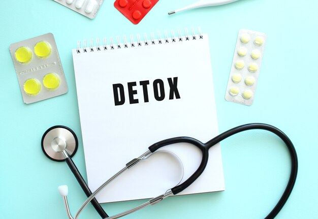 The text DETOX is written on a white notepad that lies next to the stethoscope and pills on a blue background.