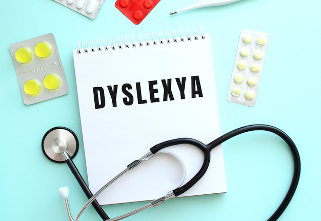 The text DYSLEXYA is written on a white notepad that lies next to the stethoscope and pills on a blue background. Medical concept